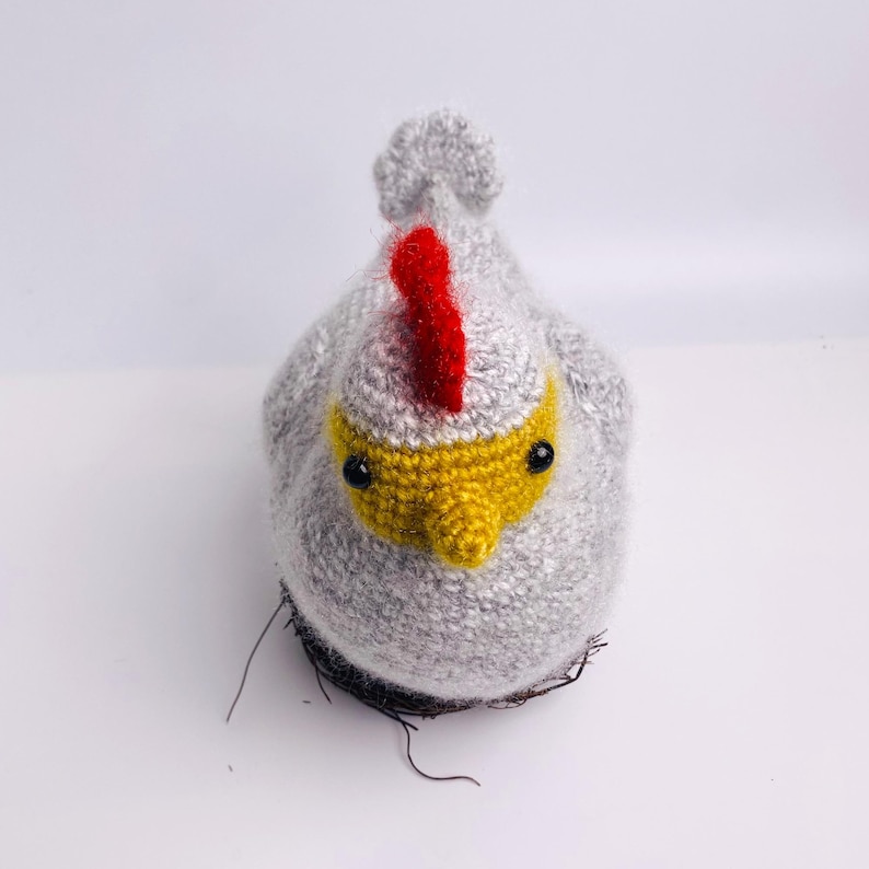 May include: A handmade, gray crocheted chicken with a red comb and yellow beak. The chicken has black eyes and is sitting on a nest made of dark brown yarn. The craft project is a whimsical decorative item.