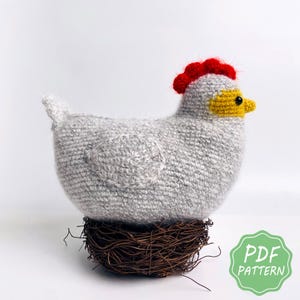 May include: A handmade gray crochet chicken with a red comb and yellow beak, perched on a brown twig nest. The chicken has a round body and a small tail. The image includes a green and white "PDF PATTERN" graphic.