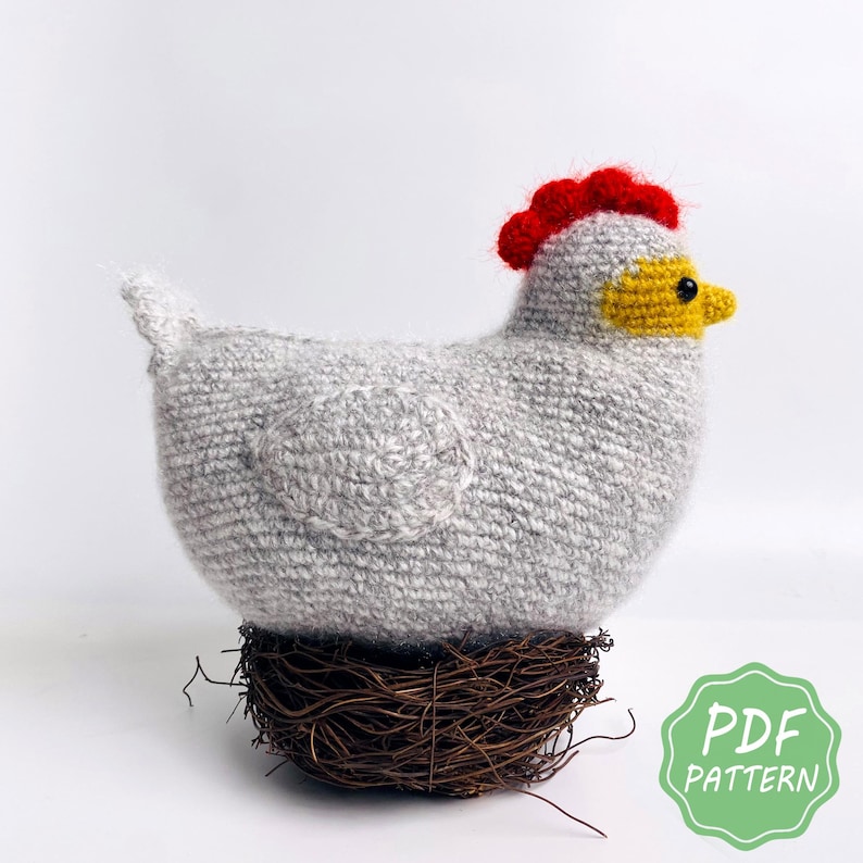 May include: A handmade gray crochet chicken with a red comb and yellow beak, perched on a brown twig nest. The chicken has a round body and a small tail. The image includes a green and white "PDF PATTERN" graphic.