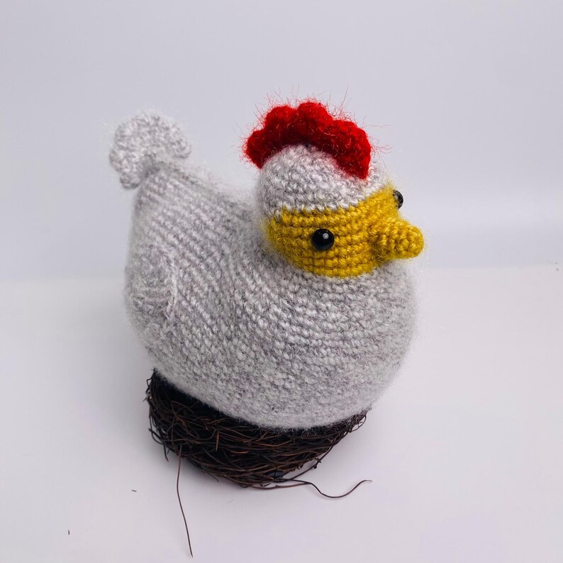 May include: A handmade gray knitted chicken with a red comb and yellow beak, perched on a small brown nest. The chicken has black eyes and a fluffy tail. This decorative item is perfect for Easter or farmhouse decor.