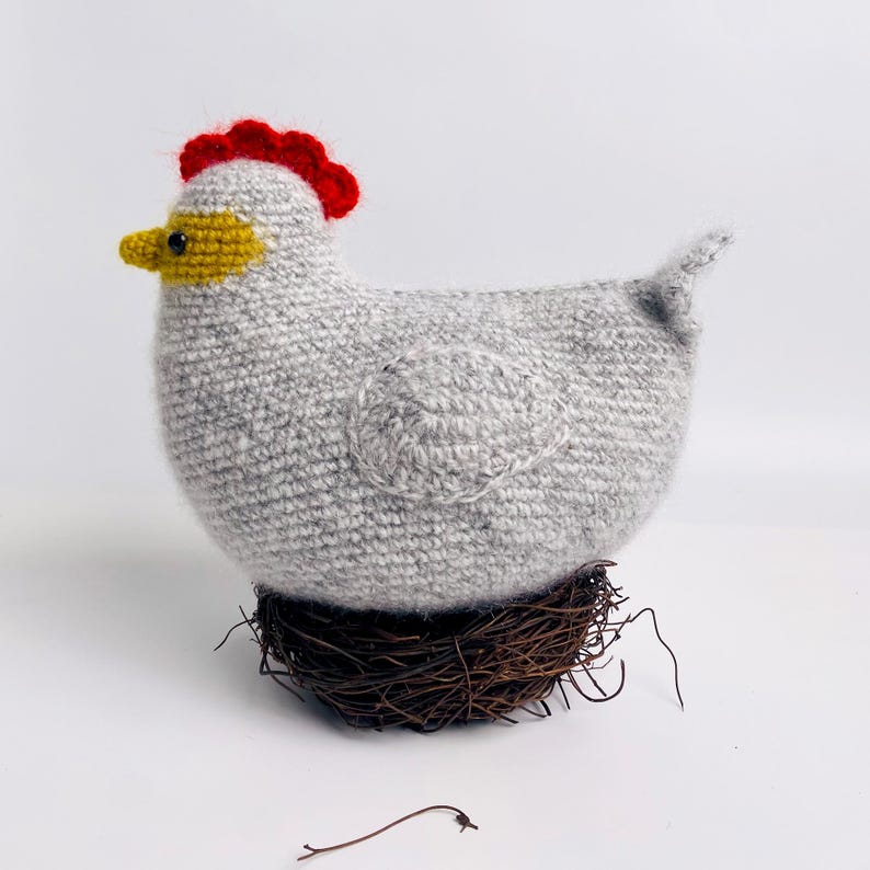 May include: A handmade, gray crocheted chicken with a red comb, yellow beak, and a brown nest. The chicken is a decorative item, possibly for Easter or farmhouse decor.