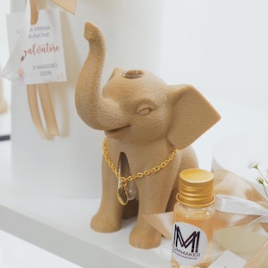 Può includere: Una statuetta di elefante marrone con una collana a catena dorata e una piccola bottiglia con tappo dorato. La statuetta è accanto a un cilindro bianco con un nastro e un'etichetta "Salvatore 3 Maggio 2026".