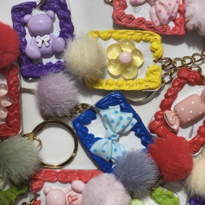 Peut inclure: Une collection de porte-clés rectangulaires avec des cadres texturés de différentes couleurs, notamment violet, jaune, bleu, rouge et vert. Chaque porte-clés est orné d'un motif central comme un ours, un nœud ou une fleur et d'un pompon.