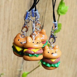 Peut inclure: Quatre breloques miniatures en forme de burger chat, avec des cordons noirs et des accessoires argentés. Les burgers ont des pains bruns, du fromage jaune, de la tomate rouge et de la laitue verte. Les breloques sont suspendues sur un fond flou.