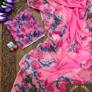 May include: A bright pink sheer fabric garment featuring a floral pattern in shades of dark blue, magenta, and white, accented with small reflective sequins. A pair of matching earrings with red beads and gold-tone metal accents is included.