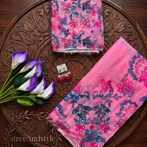 May include: A pink fabric piece with a floral pattern in shades of dark blue and magenta, accented with silver sequins. A pair of silver-toned earrings with red beads is included. The fabric features a repeating botanical print throughout.