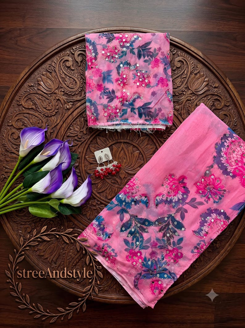 May include: A pink fabric piece with a floral pattern in shades of dark blue and magenta, accented with silver sequins. A pair of silver-toned earrings with red beads is included. The fabric features a repeating botanical print throughout.