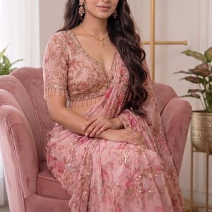 May include: A light pink saree featuring intricate floral embroidery with sequins and beads. The blouse has a deep V-neckline, short sleeves with beaded fringe trim, and matching floral patterns. The logo streeAndstyle Authentic Handwork is visible.