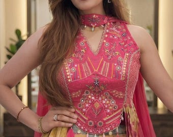 May include: A sleeveless pink bodice featuring intricate embroidery, mirror work, and a high neck collar with a matching choker. The garment includes a draped, multi-colored skirt with yellow, grey, and pink patterns, accented by hanging tassels.