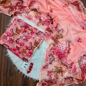May include: A peach-colored fabric with a floral pattern in shades of pink, red, and brown, accented with small circular mirrors and sequins. The set includes a folded piece of the same fabric and a pair of matching dangling earrings with red beads.