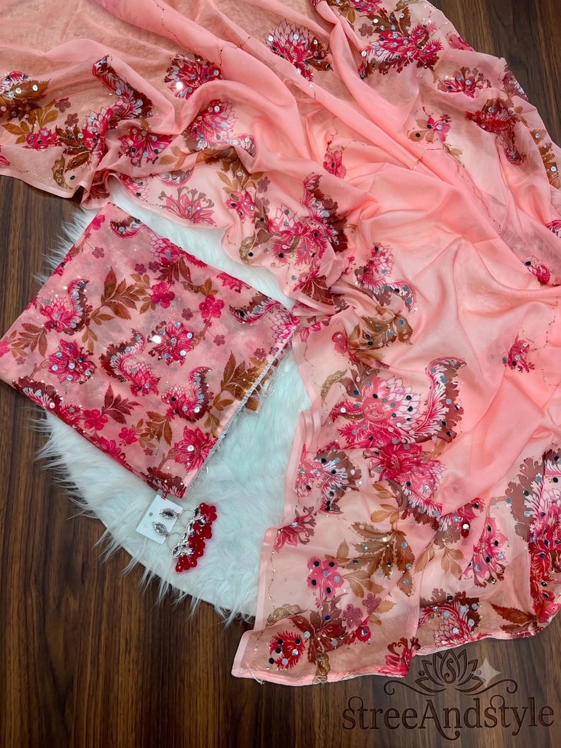 May include: A peach-colored fabric with a floral pattern in shades of pink, red, and brown, accented with small circular mirrors and sequins. The set includes a folded piece of the same fabric and a pair of matching dangling earrings with red beads.