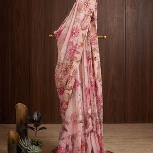 May include: A light pink saree featuring a floral print in shades of dark pink and brown, accented with gold embroidery and sequins. The matching blouse has a deep V-neckline, short sleeves with beaded fringe, and intricate gold embellishments.