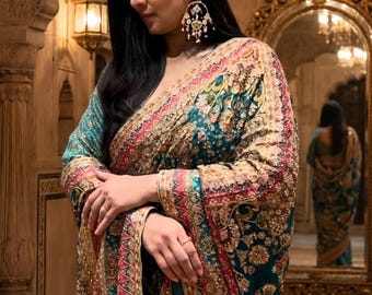 May include: A teal saree featuring intricate gold embroidery, floral patterns, and a wide border with red and gold geometric detailing. The garment is draped over the shoulder and includes ornate, dangling earrings with pink and white accents.