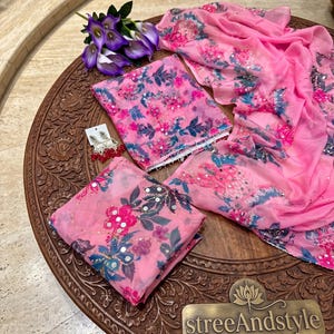 May include: A pink fabric set featuring a floral pattern in shades of blue, dark green, and magenta, accented with small reflective mirror embellishments. The set includes a folded top, a matching dupatta, and a pair of dangling earrings.