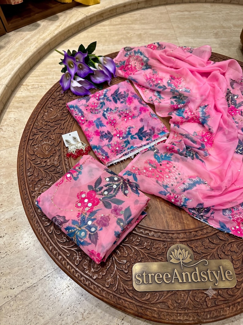 May include: A pink fabric set featuring a floral pattern in shades of blue, dark green, and magenta, accented with small reflective mirror embellishments. The set includes a folded top, a matching dupatta, and a pair of dangling earrings.