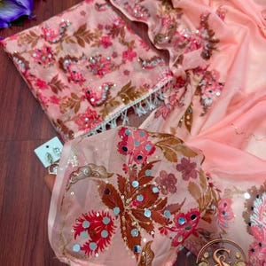 May include: A peach-colored fabric featuring a floral pattern in shades of pink, brown, and white, embellished with small circular mirrors and sequins. The material has a sheer texture and includes a matching folded piece with white fringe trim.