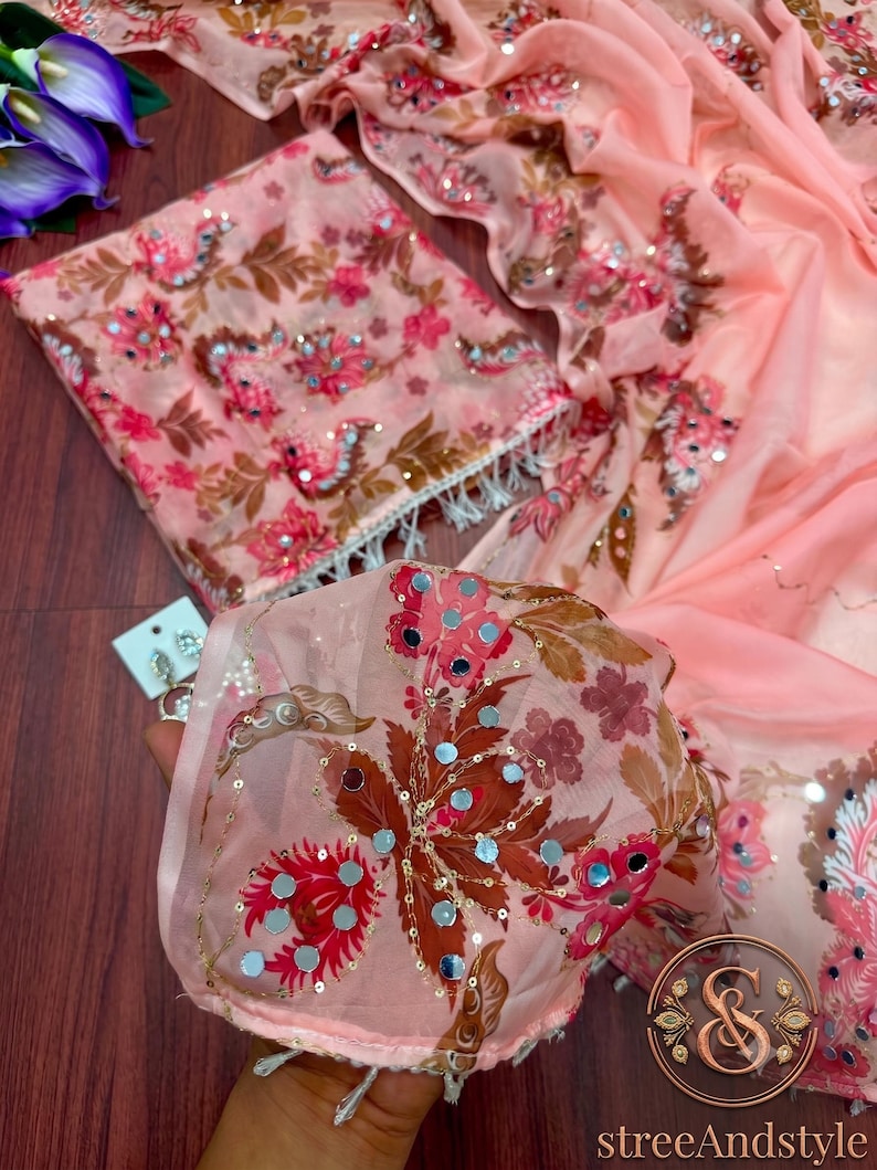 May include: A peach-colored fabric featuring a floral pattern in shades of pink, brown, and white, embellished with small circular mirrors and sequins. The material has a sheer texture and includes a matching folded piece with white fringe trim.