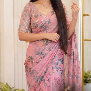 May include: A light pink saree featuring a floral print in shades of blue and green, accented with small circular mirrors. The garment includes a matching blouse with short sleeves and mirror work along the neckline and edges. Text reads streeAndstyle.