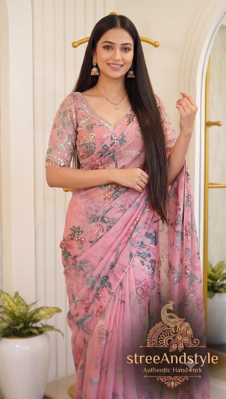 May include: A light pink saree featuring a floral print in shades of blue and green, accented with small circular mirrors. The garment includes a matching blouse with short sleeves and mirror work along the neckline and edges. Text reads streeAndstyle.