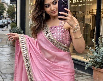 May include: A light pink saree featuring a sleeveless blouse with a square neckline and a dense pattern of small square mirrors. The saree drape includes a wide border embellished with rectangular mirrors and gold trim, creating a reflective finish.