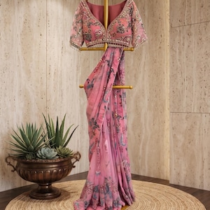 May include: A pink saree featuring a floral print and intricate embroidery on the blouse. The garment includes a matching pallu with decorative borders and embellishments. A small sign at the base displays the text streeAndstyle in gold lettering.
