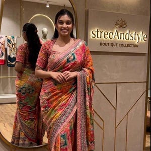 May include: A woman wearing a colorful saree with a floral and geometric pattern. The saree features shades of pink, orange, and green. The image also shows a mirror and a sign that says "streeAndstyle BOUTIQUE COLLECTION."