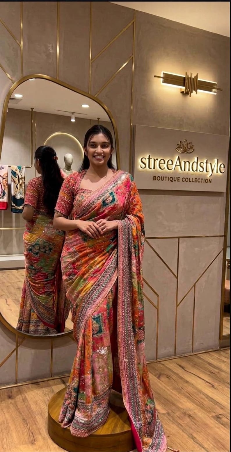 May include: A woman wearing a colorful saree with a floral and geometric pattern. The saree features shades of pink, orange, and green. The image also shows a mirror and a sign that says "streeAndstyle BOUTIQUE COLLECTION."
