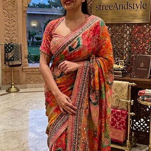 May include: A vibrant, patterned saree in shades of red, orange, and green. The saree features intricate embroidery and embellishments. The image also displays the text "streeAndstyle".