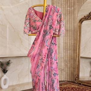 May include: A pink saree featuring a floral print in shades of blue, grey, and white, accented with intricate silver embroidery, sequins, and beadwork along the borders and blouse, displayed on a gold-toned stand with the text streeAndstyle.