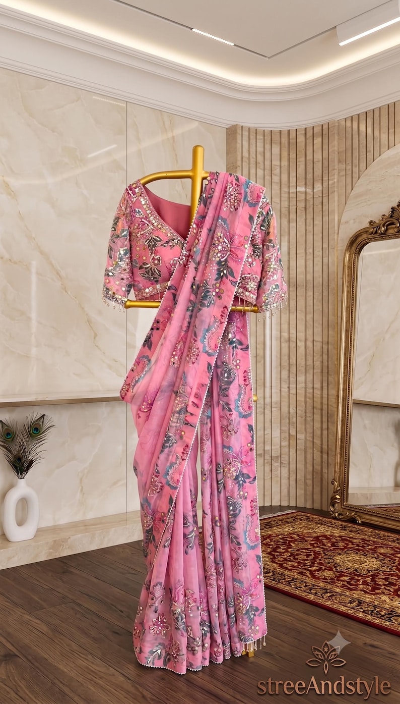 May include: A pink saree featuring a floral print in shades of blue, grey, and white, accented with intricate silver embroidery, sequins, and beadwork along the borders and blouse, displayed on a gold-toned stand with the text streeAndstyle.