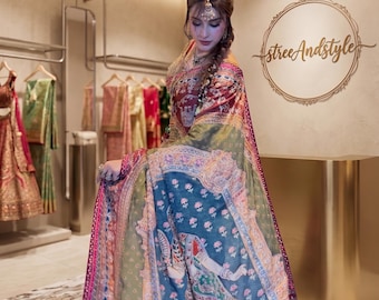 May include: A multi-colored saree featuring intricate patterns in shades of blue, green, red, and gold. The fabric displays detailed illustrations of figures and floral motifs, with a prominent border and a draped silhouette worn over the shoulder.