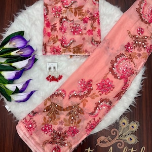 May include: A peach-colored fabric set featuring a floral pattern with brown and pink tones, accented by small reflective mirror embellishments. The set includes a folded piece of fabric, a long draped length, and a pair of dangling earrings.