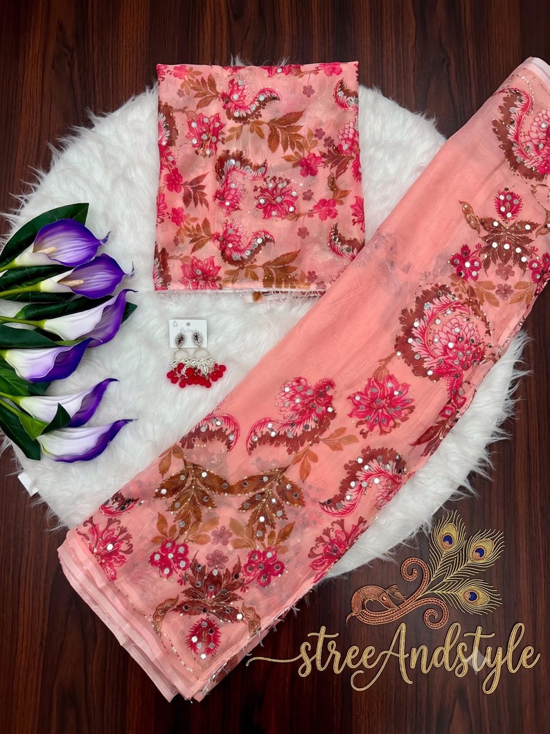 May include: A peach-colored fabric set featuring a floral pattern with brown and pink tones, accented by small reflective mirror embellishments. The set includes a folded piece of fabric, a long draped length, and a pair of dangling earrings.