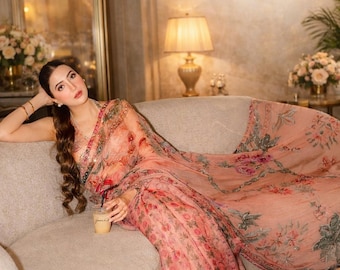 May include: A peach-colored saree featuring a delicate floral print in shades of red, pink, and green. The fabric is sheer and lightweight, adorned with intricate embroidery along the borders. The garment is draped elegantly over the shoulder.