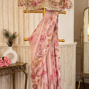 May include: A light pink saree featuring a floral pattern in shades of pink, brown, and gold, with intricate embroidery, sequins, and mirror work. The matching blouse has short sleeves, a V-neckline, and decorative beaded trim along the hem.
