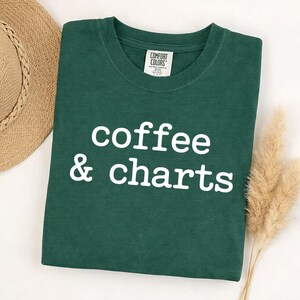 May include: A dark green t-shirt with the words "coffee & charts" in white block letters. The shirt is folded and has a tag on the collar. A straw hat and dried ornamental grass are in the background.