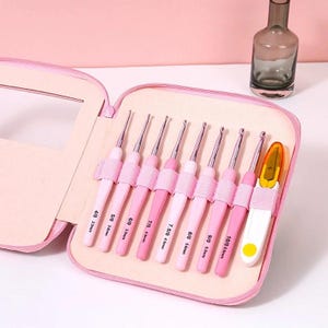 May include: A set of eight crochet hooks with pink handles and silver metal tips, ranging from 2.5mm to 6.0mm, organized in a pink zippered case alongside a white and yellow seam ripper with a clear plastic cap.
