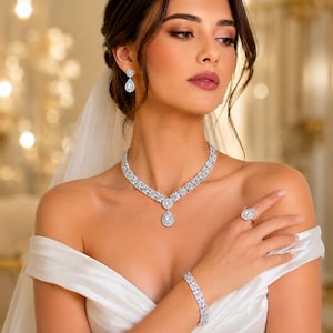 May include: A silver-toned jewelry set featuring a necklace with a pear-shaped pendant, matching drop earrings, a tennis bracelet, and a ring. Each piece is encrusted with clear, sparkling stones arranged in floral-inspired clusters.