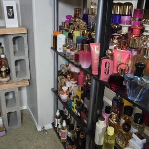 May include: A black shelving unit filled with various perfume bottles of different shapes, sizes, and colors. Some bottles have gold accents. The shelf is against a white wall and a beige carpeted floor. A small pink computer mouse is on the floor.