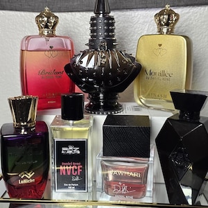 May include: An assortment of perfume bottles with unique designs. The bottles vary in shape, color, and size, with gold, black, and clear glass. Some bottles have crown-shaped tops, while others feature geometric designs. The text "Brilant", "Mouillée", "NVCF", and "JAWHARI" is visible.