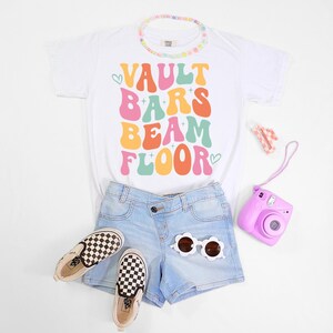 Puede incluir: Una camiseta blanca con las palabras "VAULT BARS BEAM FLOOR" en una fuente retro colorida. La imagen incluye pantalones cortos de mezclilla azul claro, zapatillas sin cordones a cuadros, un collar de cuentas, gafas de sol de flores, una cámara rosa y una pinza para el pelo.