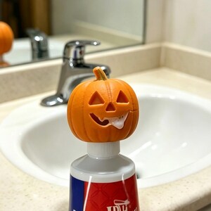 May include: A Halloween-themed orange pumpkin-shaped toothpaste cap with a carved face, sitting atop a tube of toothpaste. The toothpaste appears to be squeezed out of the pumpkin's mouth. The background includes a bathroom sink and mirror.