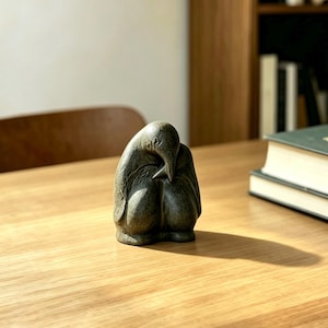 Può includere: Una scultura in pietra verde scuro raffigurante un pinguino in posizione rannicchiata e a riposo. La figura presenta una texture liscia e opaca con sottili linee incise che indicano ali e piumaggio, creando una forma arrotondata e compatta.