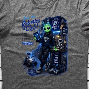 May include: A heather gray t-shirt featuring a graphic of a green alien wearing a black hoodie and sneakers, standing in a workshop. The text reads Join the Robotics Club, Build, Learn, Innovate, Collaborate, Shape the Future, Your Ideas Our Future, Think Code Build Repeat.