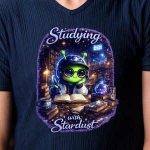 May include: Navy blue t-shirt with a cartoon alien studying. The alien is wearing a helmet and glasses. The text "Studying with Stardust" is displayed in a purple, starry design. The image includes books, a cup, and an hourglass.
