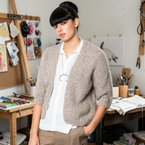Könnte beinhalten: Eine beige, strukturierte Strickjacke mit Dreiviertelärmeln und offener Vorderseite, getragen über einem weißen Hemd mit Knopfleiste. Eine dünne silberne Halskette mit einem großen, kreisförmigen Anhänger wird zu einer braunen Hose kombiniert.