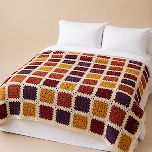 Può includere: Una coperta all'uncinetto fatta a mano con un design patchwork. La coperta presenta quadrati nei toni dell'arancione, del rosso, del giallo e del viola, bordati da un bordo color crema. La coperta è esposta su un letto bianco con cuscini bianchi.