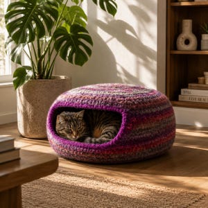 May include: A cozy, handmade cat bed in shades of purple, pink, and brown. The crocheted bed has a rounded shape with a rectangular opening, and a tabby cat is nestled inside. The bed is on a wooden floor, with a large potted plant in the background.