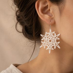 May include: White, snowflake-shaped crochet earrings. The earrings are made of a delicate, open-weave crochet pattern, with a central circular design and radiating points. The earrings hang from silver-colored hooks.