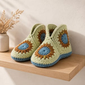 Op de afbeelding: Een paar handgemaakte gehaakte pantoffels in een granny square design. De pantoffels hebben een lichtgroene basis met blauwe, bruine en gele bloemaccenten. De zolen en de rand zijn blauw. De pantoffels worden op een houten plank getoond.